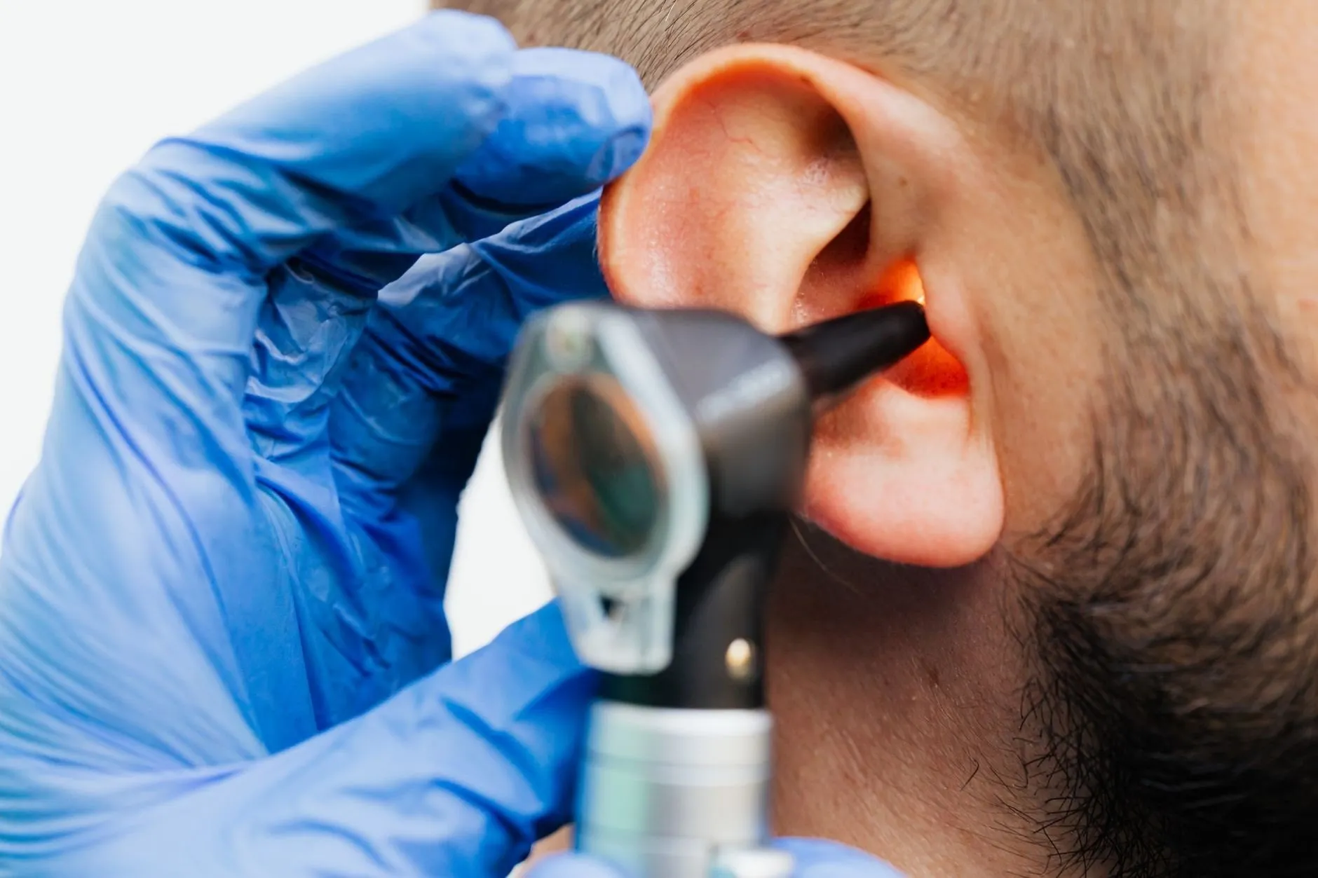 A gloved audiologist looking into a patient's ear with an otoscope, the ear canal lit orange
