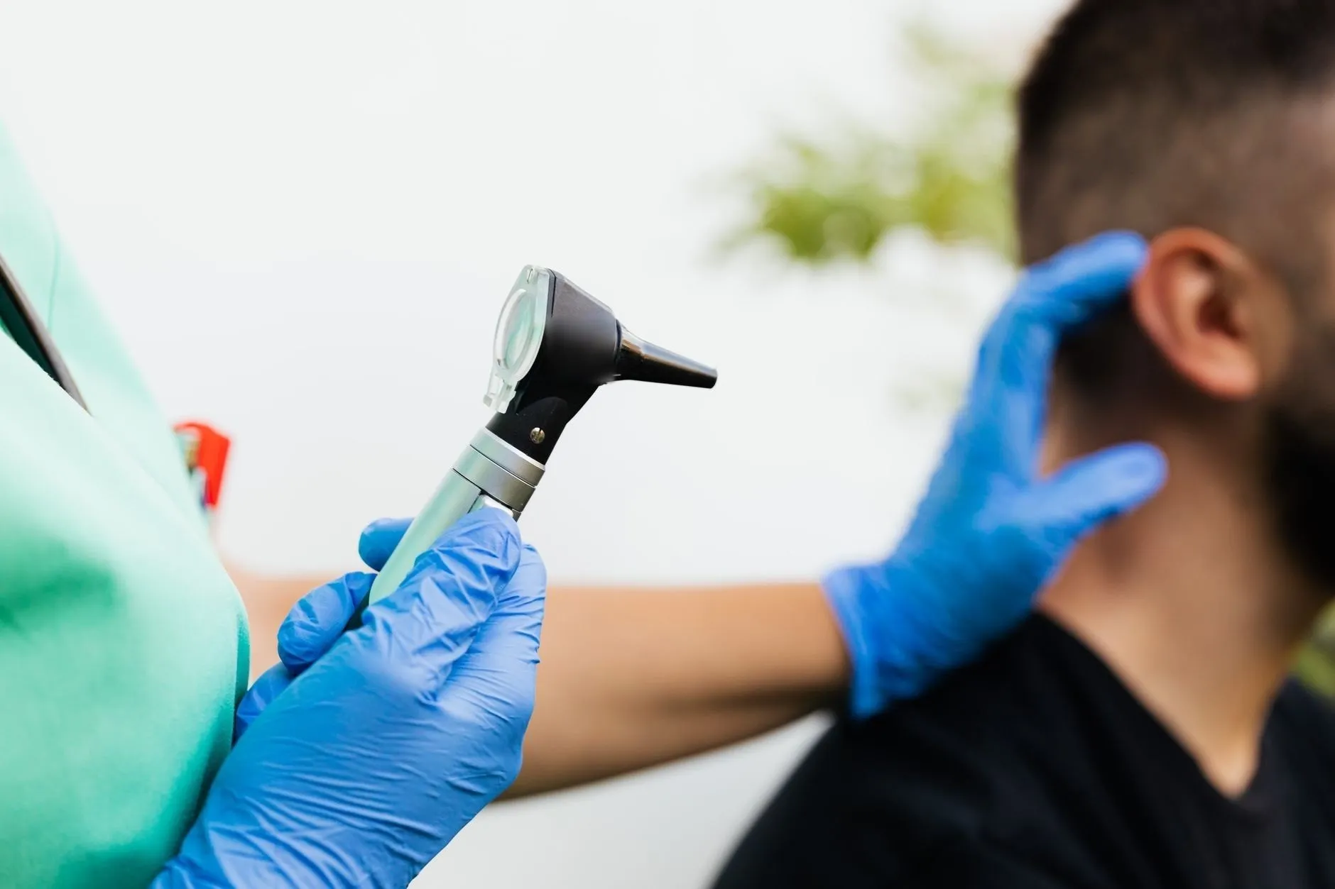 An audiologist in blue gloves holding an otoscope beside a seated patient's ear